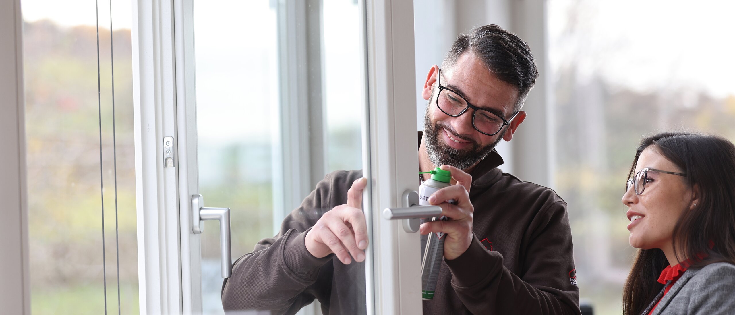 Ein Mann und eine Frau vor einem offenen Fensterflügel. Der Mann sprüht mit einer Flasche auf den Beschlag des Fensterflügels