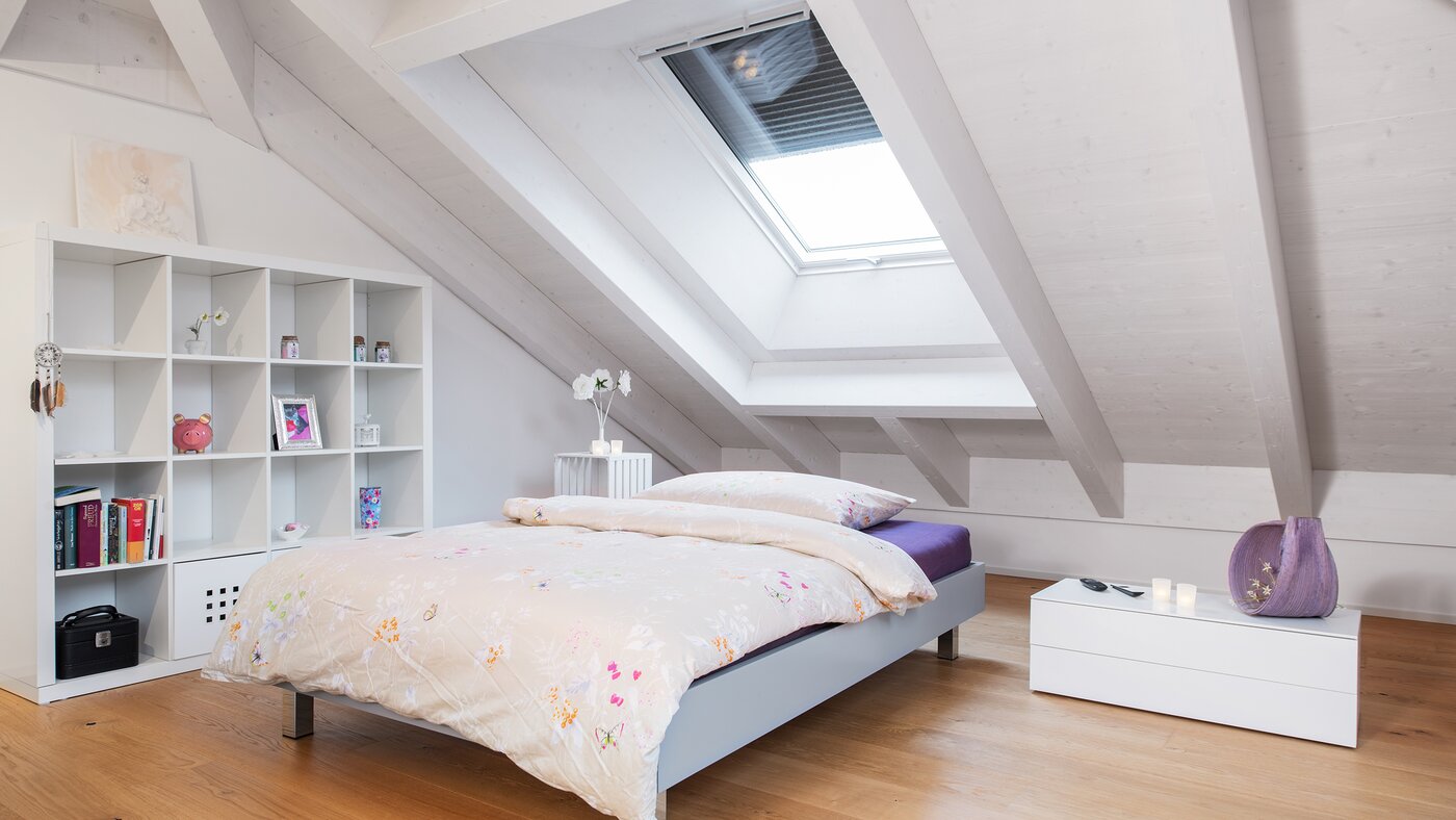 Ein Schlafzimmer mit einem Dachfenster. Das Dachfenster hat ein Rollo welches zur hälfte geschlossen ist.