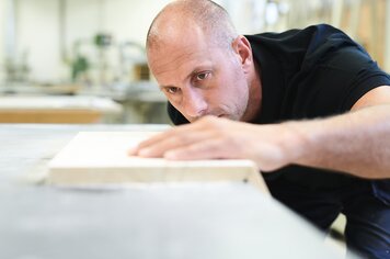 Ein Mann in einer Werkstatt. Er hat seine Hand auf einem Holzbrett und konzentriert darauf schaut.
