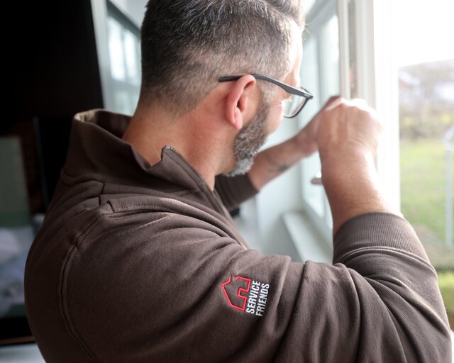 Ein Mann mit Service Friends Pullover. Er schraubt an der Innenseite eines Fensterflügels. 