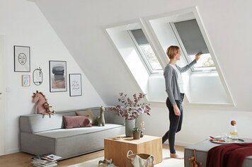 Ein Raum mit zwei Dachfenstern. Eine Frau, die an einem Fenster das Rollo für Sicht und Lichtschutz herunterzieht. 