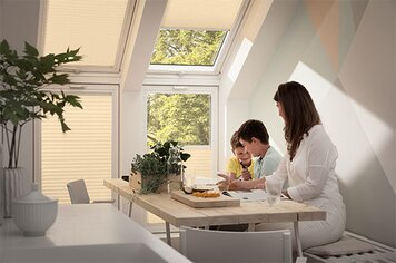 Ein Raum in dem eine Frau mit zwei Kindern auf Stühlen sitzt. Man sieht vier Dachfenster mit Faltrollos.