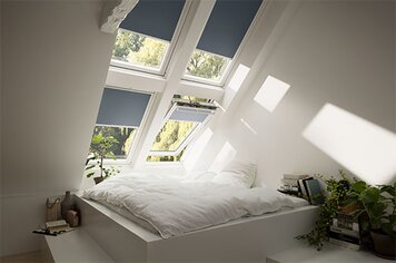 Vier Dachfenster in einem Zimmer mit Verdunkelungsrollos. Eines der unteren Dachfenster ist geöffnet.